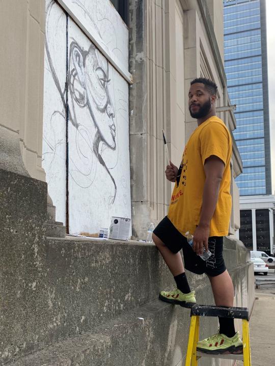 Artist Matthew Cooper looks at the camera from a step ladder in front of a sketch of a Black woman on plywood covering the old city hall