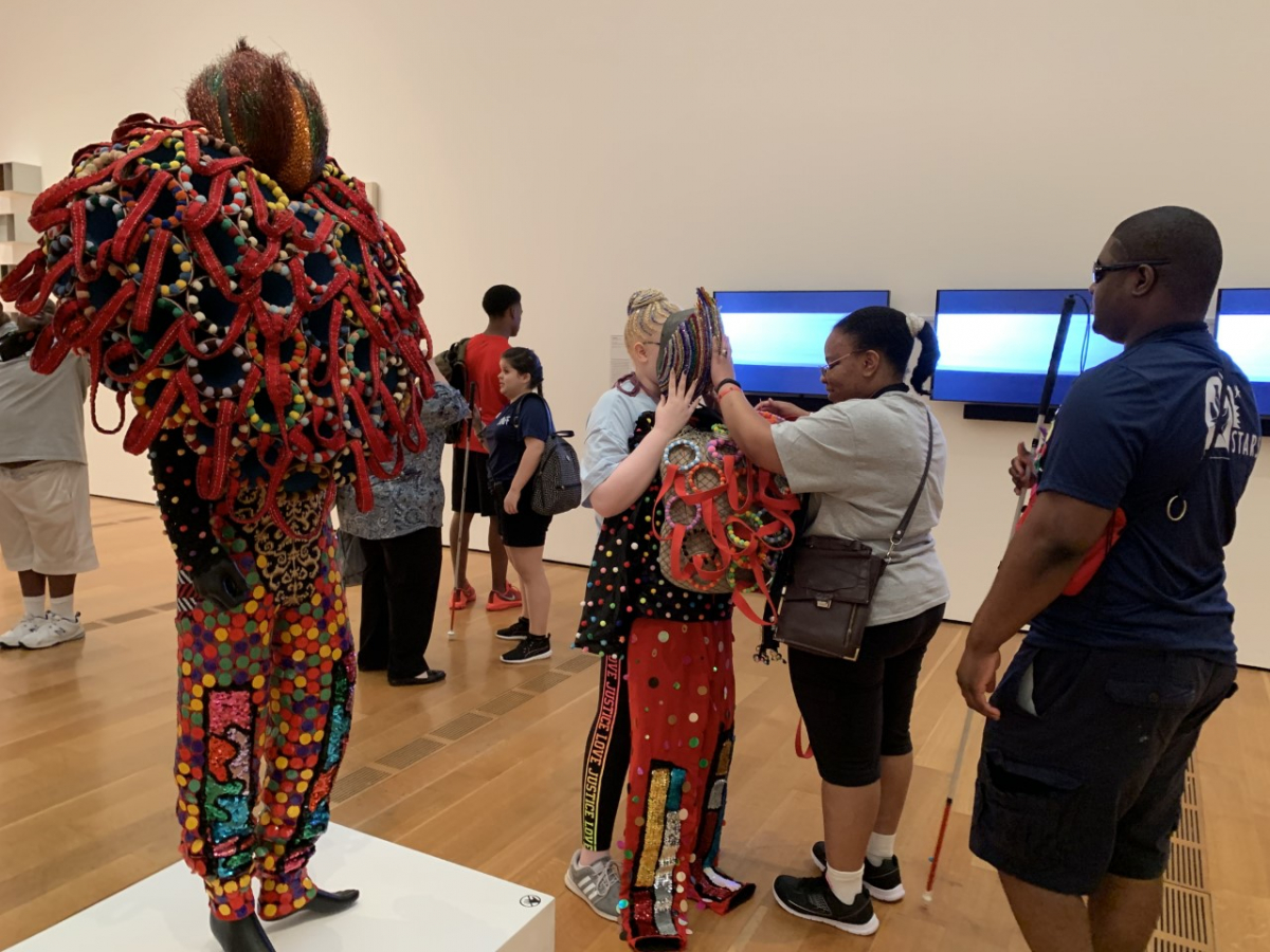 A STARS student uses her hands to feel a specially-made recreation of artwork. The original artwork, a suit of clothes covered in colorful small orbs, loops of fabric, and other textured shapes, is on display next to her in a gallery of the High Museum.