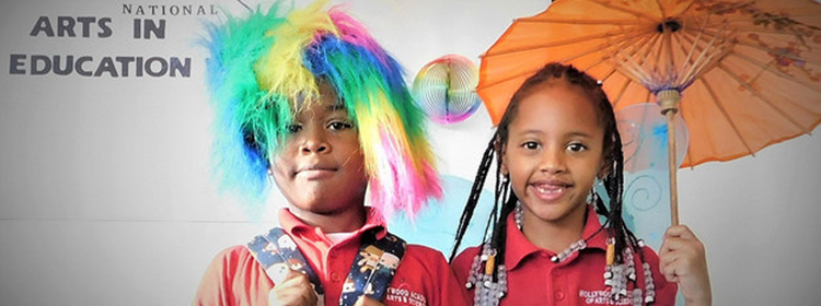 Photo of two young children (one with a colorful wig on, another with an umbrella) smiling at an event