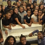 A group of students pose for the camera around a white textured piano.