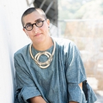 A headshot of Tanya Aguiñiga, which shows a person with close-shaved hair, wearing glasses, a large hooped necklace, and a cloudy-blue shirt.