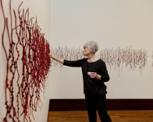 Luzene Hill, an Indigenous woman with silver hair wearing black top and pants, hangs red knotted strings on a white wall.