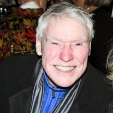 A smiling man with white hair wearing a black suit coat.